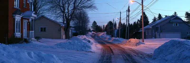 Rue résidentielle québécoise déneigée à l’aube bleutée, bancs de neige des deux côtés et lampadaire encore allumé